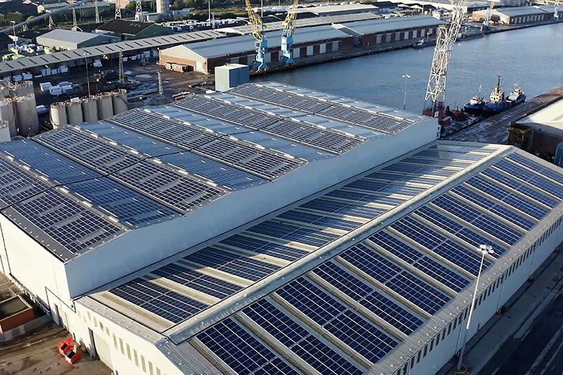 从高处俯瞰仓库的屋顶，屋顶上覆盖着蓝色的太阳能电池板. 仓库位于河边的工业区